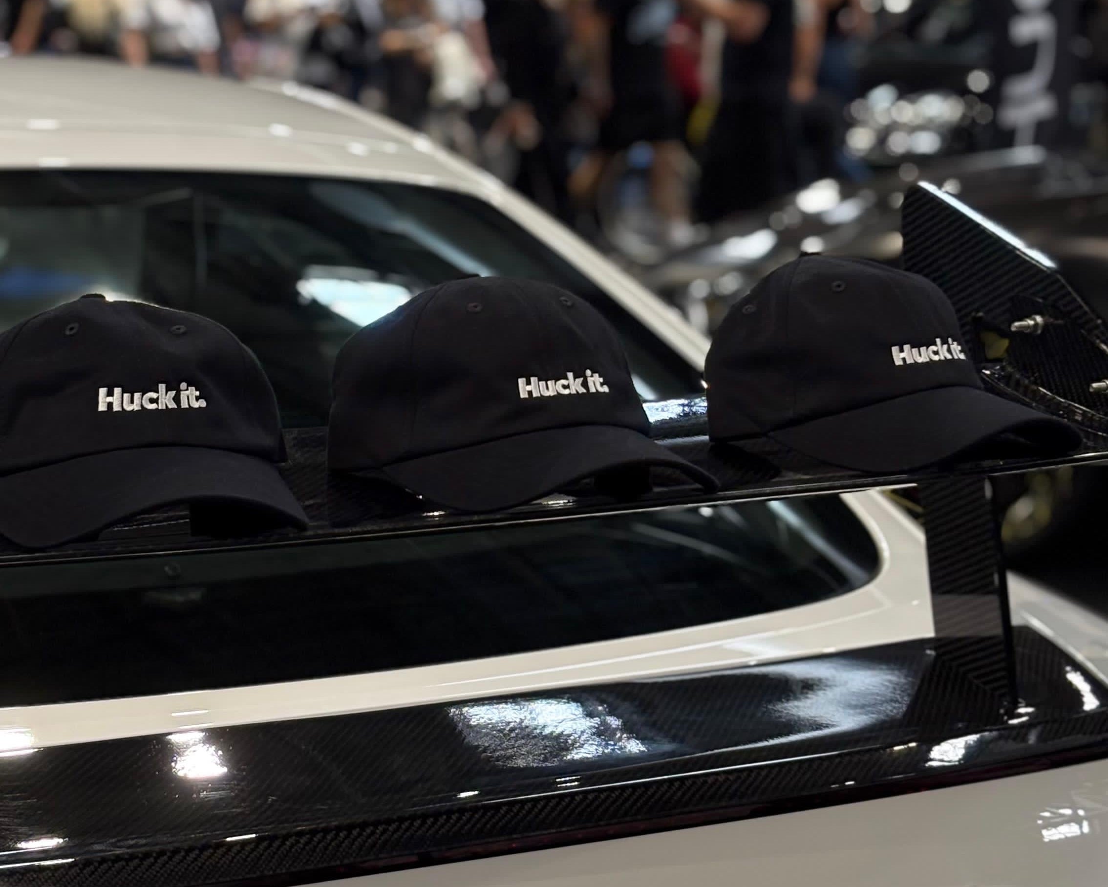 Three black baseball caps with 'Huck it.' printed in white on the front are lined up on the rear spoiler of a white Audi car at gravity show.