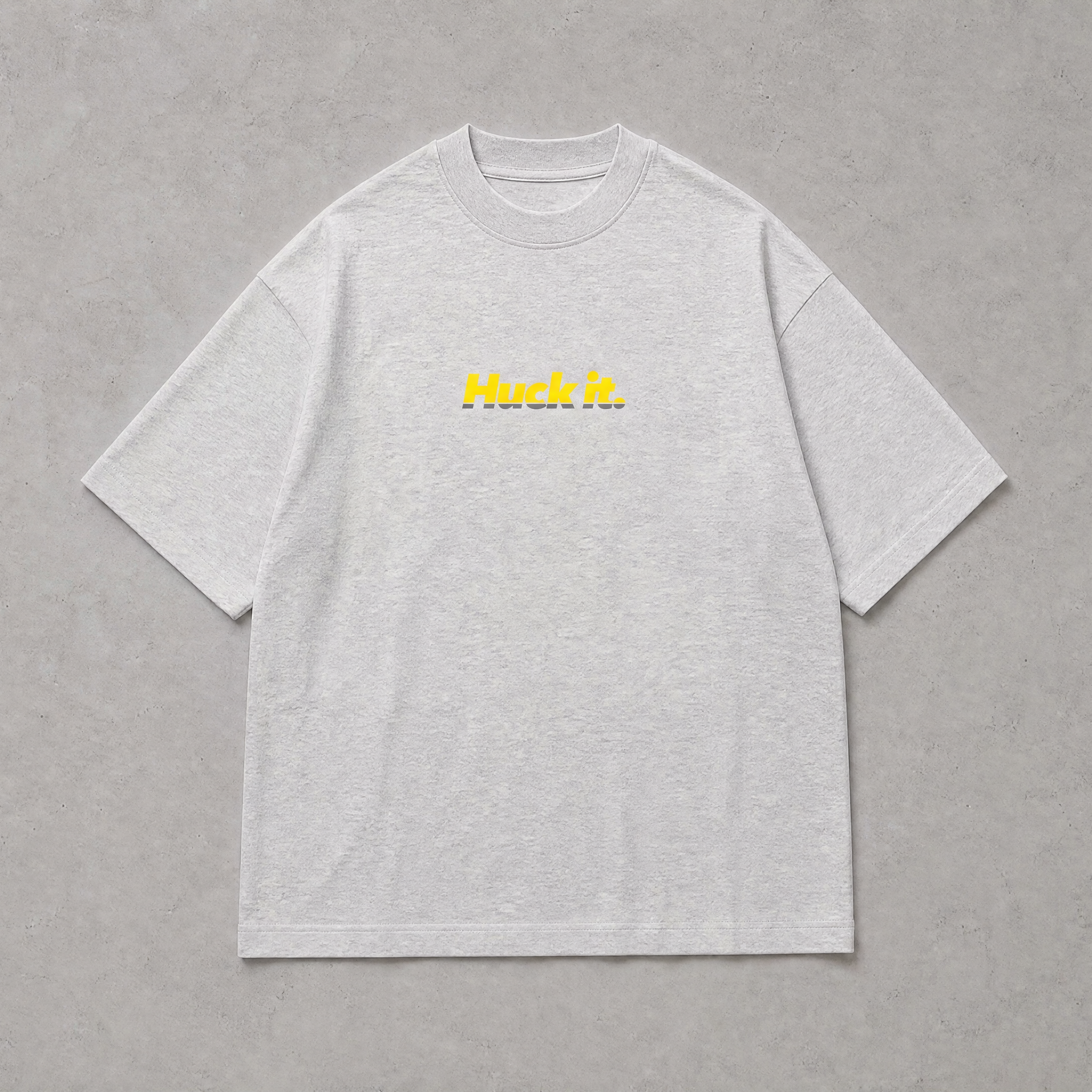 A light gray heather crewneck t-shirt with 'Huck It.' printed in yellow across the chest, displayed flat against a textured gray background.