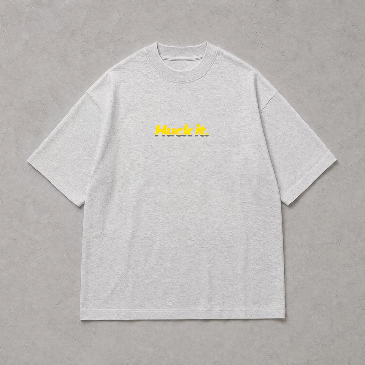 A light gray heather crewneck t-shirt with 'Huck It.' printed in yellow across the chest, displayed flat against a textured gray background.