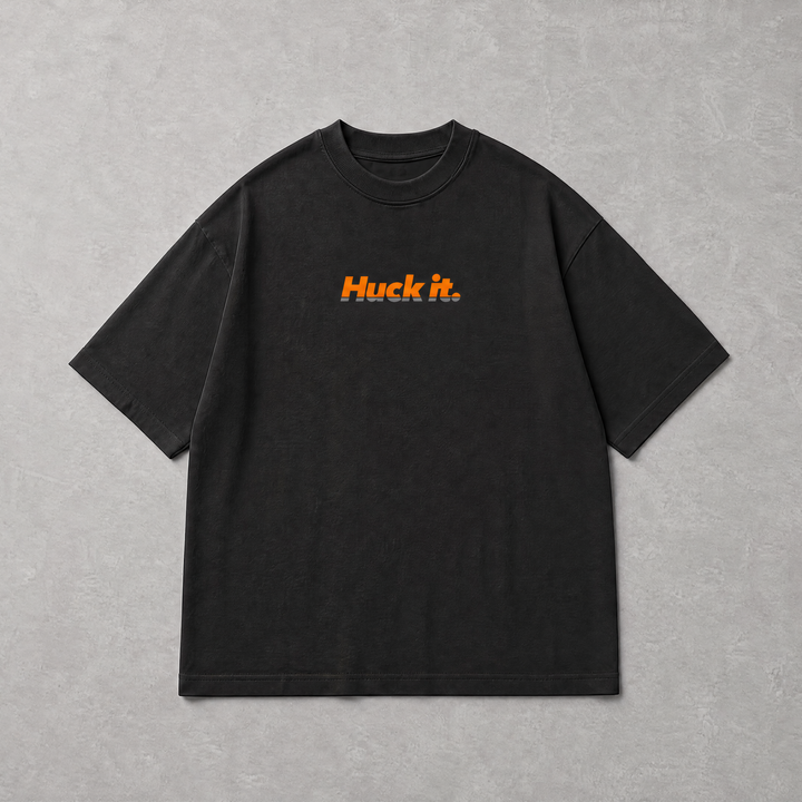 A black short-sleeve crewneck t-shirt with the orange text 'Huck It.' printed on the chest, laid flat on a textured gray background.