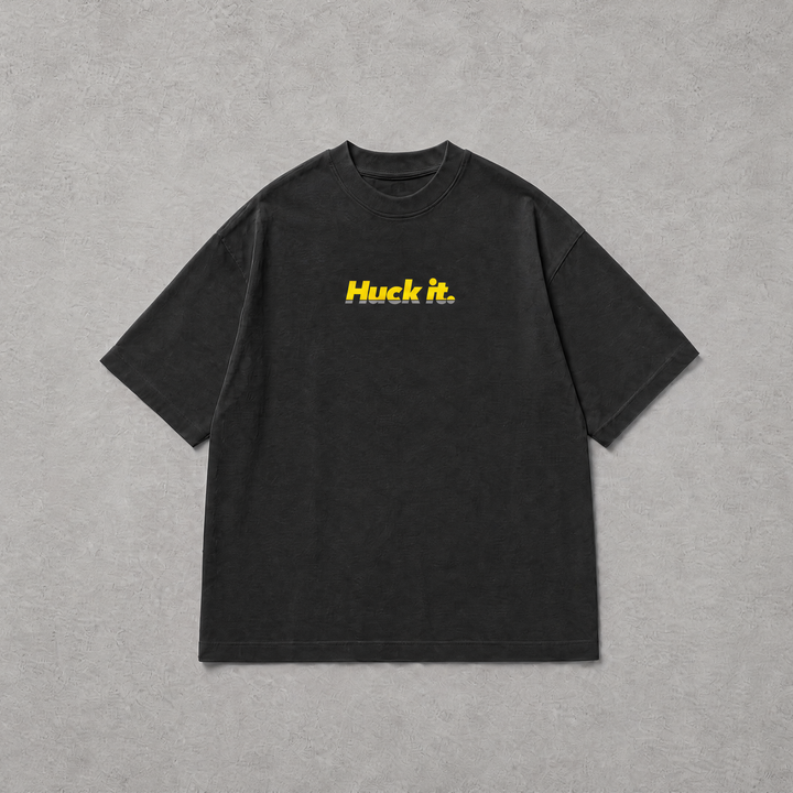 A black short-sleeve t-shirt with the text 'Huck it.' printed in yellow across the chest, laid flat on a textured gray background.