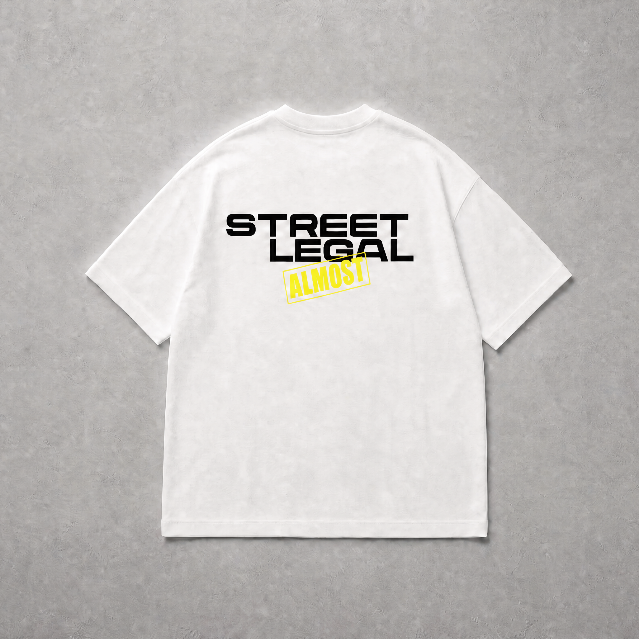 A white short-sleeve t-shirt laid flat on a textured gray background, featuring a graphic print on the back with the words 'STREET LEGAL' in black and 'ALMOST' in yellow.
